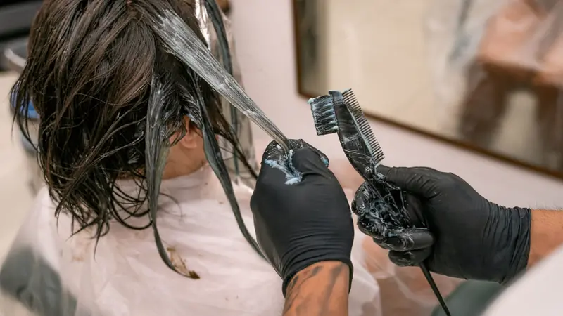 Mains gantées de noir appliquant au pinceau un produit éclaircissant bleuté sur des mèches de cheveux bruns