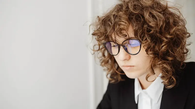 Jeune femme aux cheveux bouclés châtain clair, mi-longs et volumineux, portant des lunettes