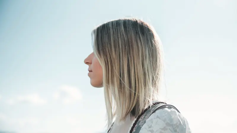 Femme aux cheveux blonds fins mi-longs, de profil, sous un ciel clair