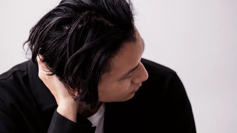 Homme aux cheveux noirs mi-longs coiffés sur le côté, la main dans les cheveux, sur fond blanc