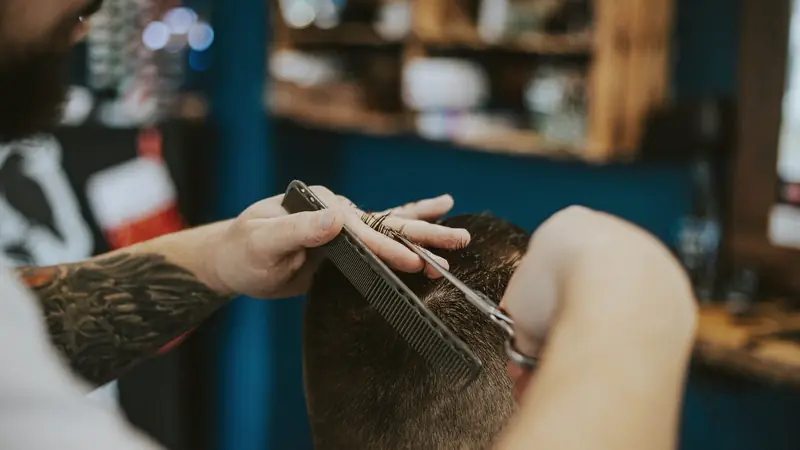 Coiffeur qui coupe aux ciseaux et au peigne le dessus de cheveux courts d'un homme vu de dos