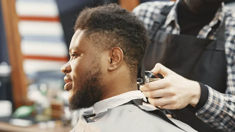 Coiffeur passant la tondeuse sur la nuque d'un homme barbu aux cheveux frisés, assis dans un fauteuil de salon