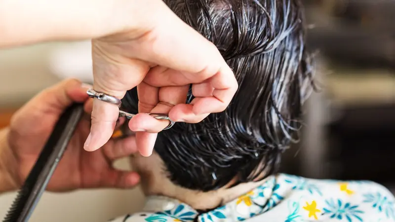 Mains d'un coiffeur tenant une mèche de cheveux courts mouillés entre deux doigts et la coupant aux ciseaux, peigne dans l'autre main