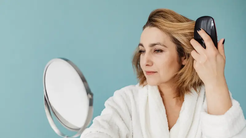 Femme en peignoir blanc aux cheveux mi-courts blonds se coiffant avec une brosse devant un miroir sur pied