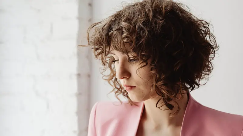 Femme aux cheveux courts bouclés châtains, coupe étagée avec frange, tête penchée devant un mur blanc