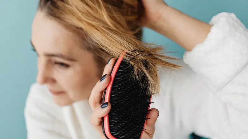Femme en peignoir démêlant doucement une mèche blonde avec une brosse souple rouge et noire