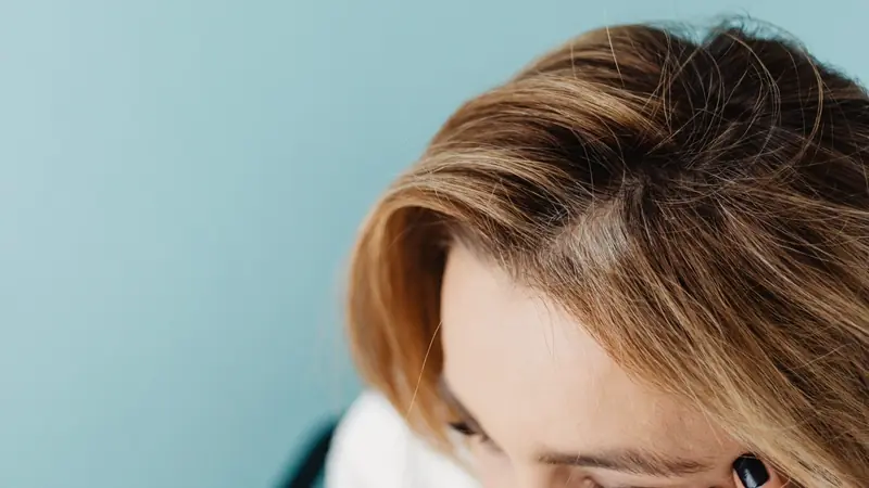 Femme aux cheveux châtain méché, tête penchée, une main au front, raie du cuir chevelu visible de près