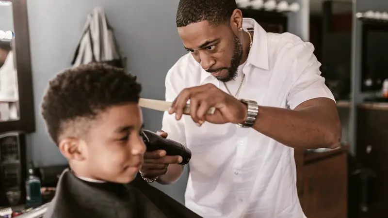 Barbier tenant un peigne et une tondeuse au-dessus de l'oreille d'un garçon aux cheveux frisés, côtés déjà dégradés