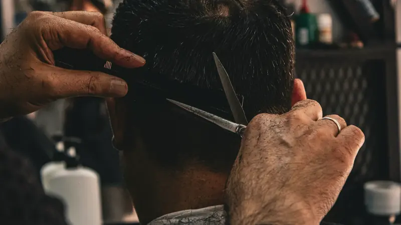 Coiffeur coupant aux ciseaux sur peigne l'arrière du crâne d'un client vu de dos, nuque courte