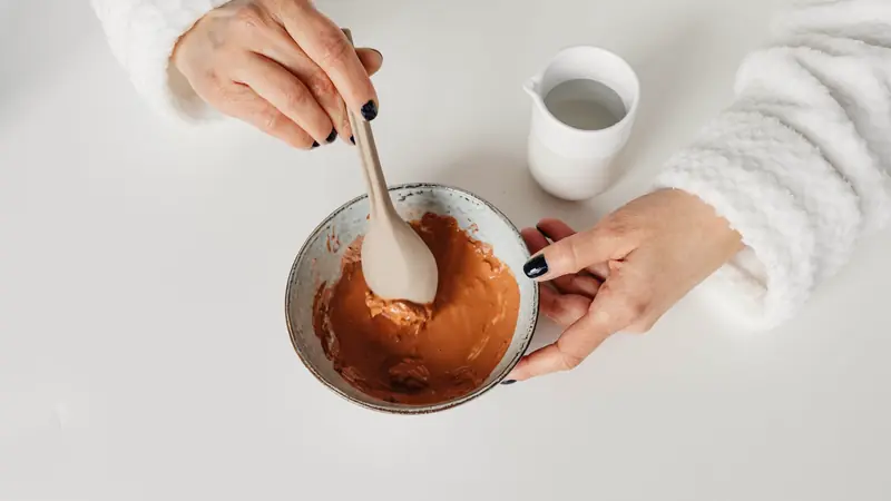 Mains mélangeant une pâte orangée dans un bol avec une cuillère, à côté d'un pot d'eau