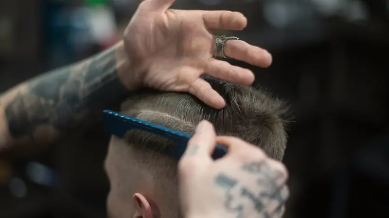 Coiffeur aux mains tatouées soulevant le dessus de cheveux courts au-dessus d'une raie et de côtés dégradés