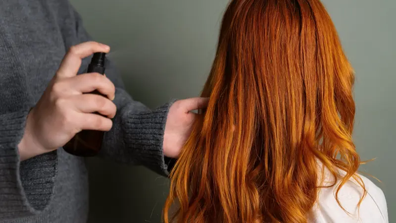 Une main vaporise un soin en spray sur de longs cheveux roux ondulés, vus de dos