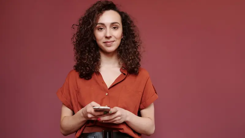 Femme aux cheveux bruns très bouclés et volumineux, téléphone en main, devant un mur rouge