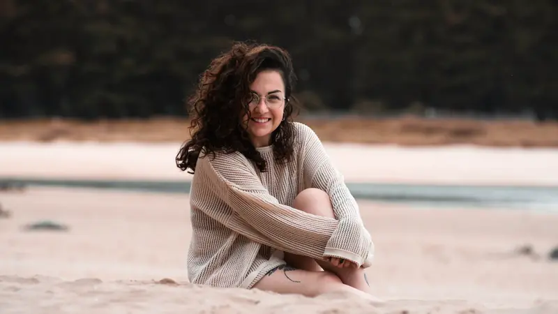 Jeune femme aux cheveux bruns bouclés et lunettes rondes, assise sur une plage de sable
