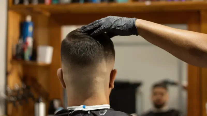 Client vu de dos chez un barbier, côtés et nuque dégradés haut jusqu'à la peau, main gantée sur le dessus