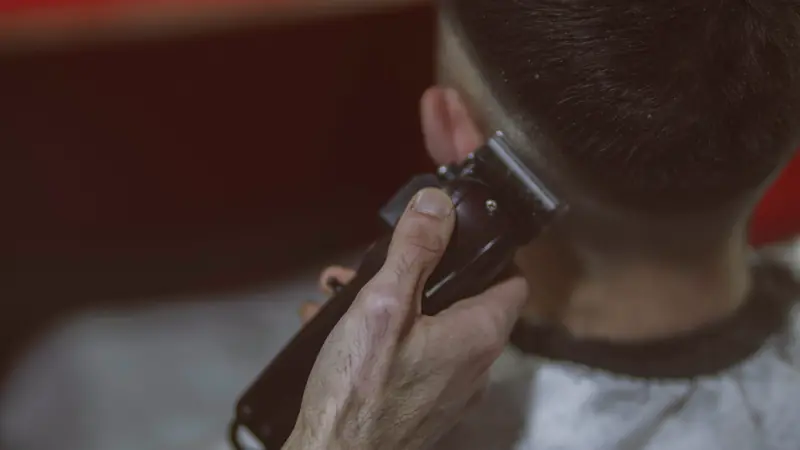Main d'un coiffeur passant une tondeuse au-dessus de l'oreille d'un client vu de dos, cheveux courts sur les côtés