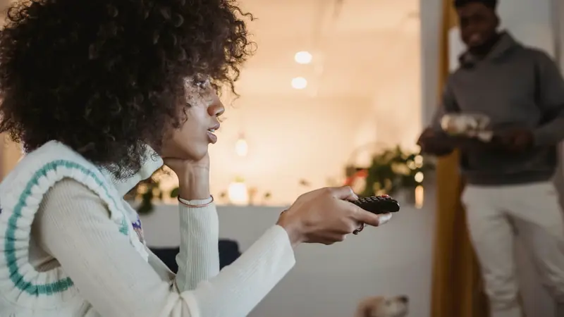 Femme aux cheveux bouclés, télécommande en main devant la télévision, pendant qu'une personne arrive avec un plateau